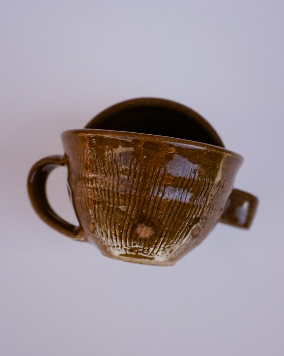 A handmade brown glazed ceramic cup with a textured vertical rib pattern, a side handle, and a small tab on the rim, shown against a plain light gray background.
