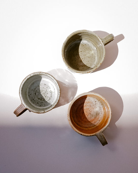 Three handmade ceramic cups with handles, shown from a top-down perspective on a light gray background with soft shadows. Each cup has a different glazed finish: one is speckled gray-green, one is speckled beige-brown, and one is speckled terracotta orange. Carved Mug | Scree Ceramics | Fodder