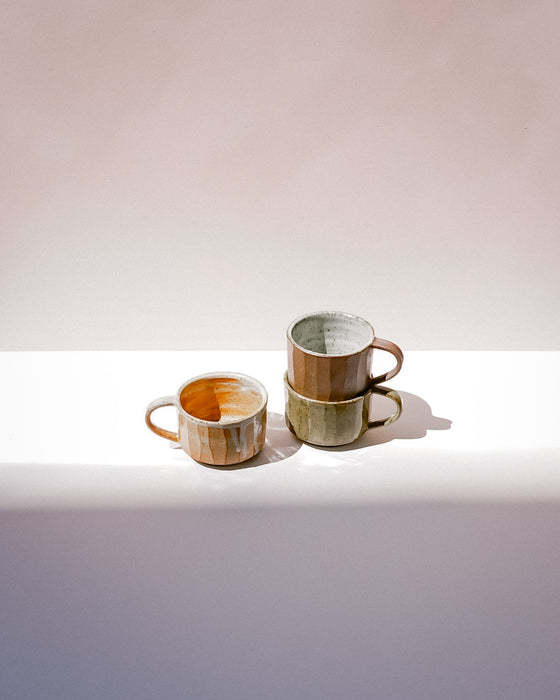 Three small ceramic mugs with a rustic, hand-painted finish in earthy tones of brown, beige, and muted green, arranged on a white surface against a warm beige background. Carved Mug | Scree Ceramics | Fodder