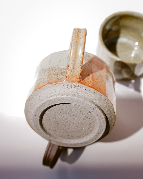 A close-up of a handmade ceramic mug showing its textured, speckled clay base and handle, with another mug blurred in the background on a white surface. Carved Mug | Scree Ceramics | Fodder