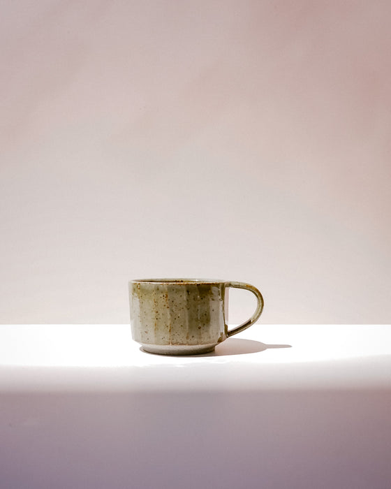 A small handmade ceramic cup with a textured, speckled glaze in earthy olive green and beige tones, sitting on a white surface against a soft, warm beige background.  Carved Mug | Scree Ceramics | Fodder