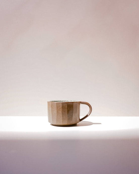 A small ceramic mug with a faceted, geometric design in a warm beige and brown glaze, sitting on a white surface against a soft beige background. Carved Mug | Scree Ceramics | Fodder