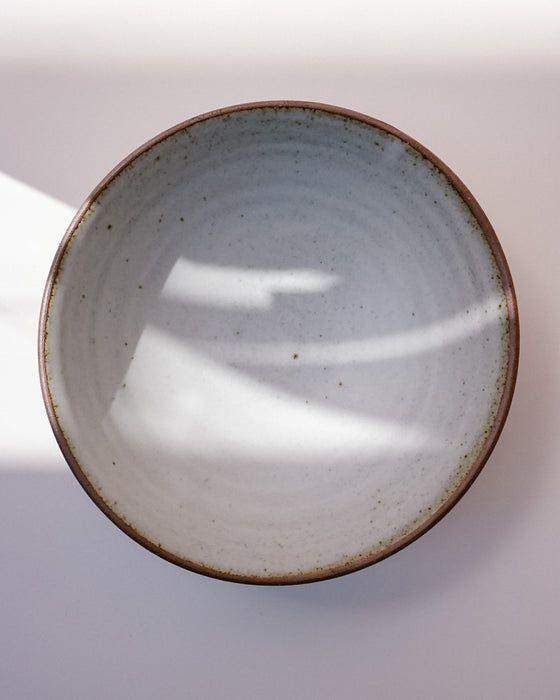 A top-down view of a handmade ceramic bowl with a speckled off-white interior and a rustic brown rim, set against a light background with soft shadows. Toasted Clay Dinner Bowl | Scree Ceramics | Fodder
