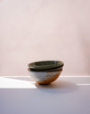 Three stacked ceramic bowls with a speckled rustic finish, featuring a dark green glazed top bowl and two natural clay bowls below, set on a white surface against a soft pink background | Scree Ceramics | Fodder