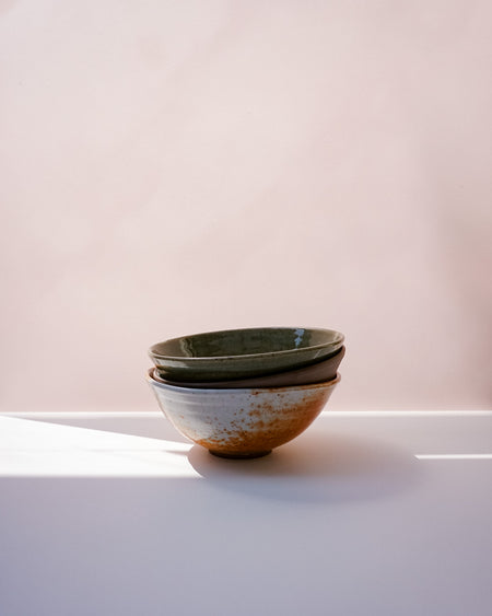 Three stacked ceramic bowls with a speckled rustic finish, featuring a dark green glazed top bowl and two natural clay bowls below, set on a white surface against a soft pink background | Scree Ceramics | Fodder