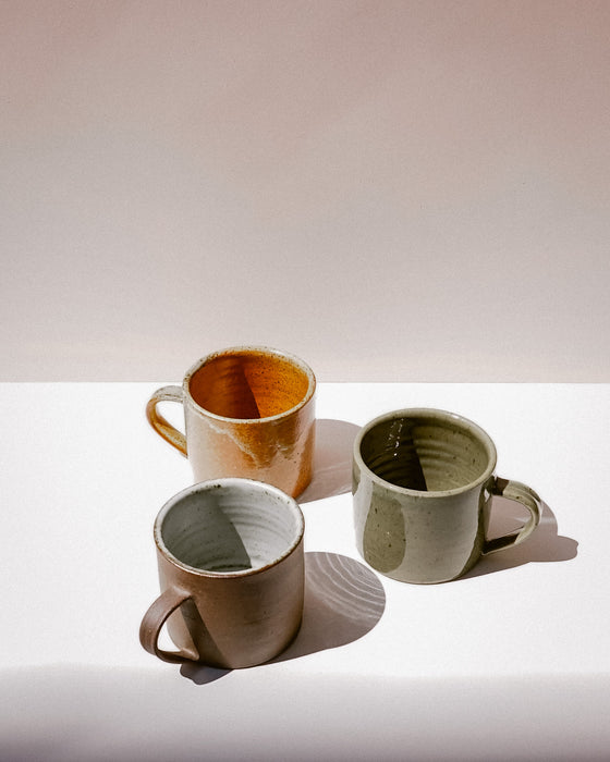 Three handmade ceramic mugs with different glazes in warm earthy tones, arranged on a white surface against a beige background. Everyday Mug | Scree Ceramics | Fodder