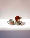 Three handmade ceramic mugs with organic shapes and handles, displayed on a white surface against a warm beige background. One mug is stacked on top of the other, showing a warm terracotta-brown interior, while the others display speckled beige and sage green exteriors. Everyday Mug | Scree Ceramics | Fodder
