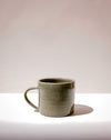 A handmade ceramic mug with a textured, speckled sage green glaze and a curved handle, sitting on a white surface against a warm beige background. Everyday Mug | Scree Ceramics | Fodder