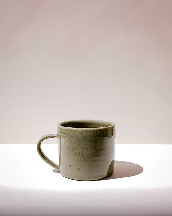 A handmade ceramic mug with a textured, speckled sage green glaze and a curved handle, sitting on a white surface against a warm beige background. Everyday Mug | Scree Ceramics | Fodder
