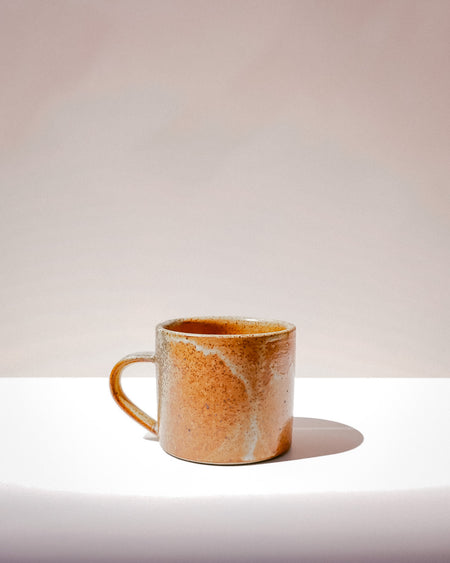 A small handmade ceramic mug with a warm orange-brown glaze and white marbling, sitting on a white surface against a light beige background. Everyday Mug | Scree Ceramics | Fodder