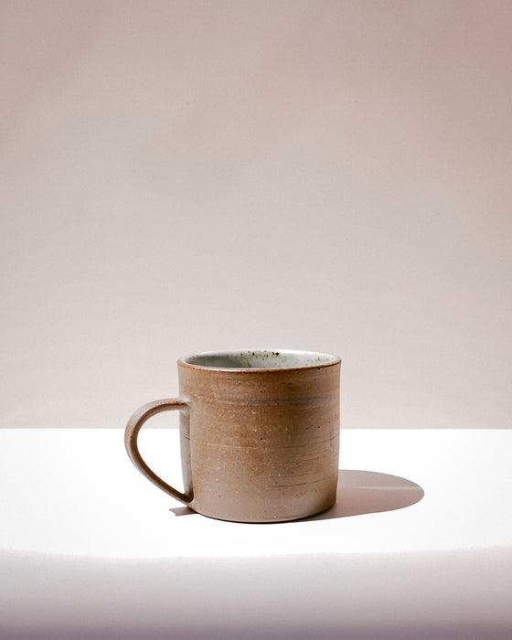 A handmade ceramic mug with a textured, earthy brown exterior and a speckled light green interior, sitting on a white surface against a warm beige background. Everyday Mug | Scree Ceramics | Fodder