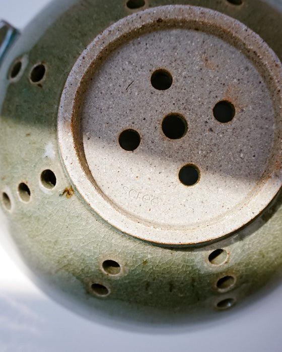 Close-up of the underside of a ceramic vessel, showing a round beige base with five drainage holes and a green glazed exterior with multiple side ventilation holes. handmade ceramic colander by Scree Ceramics | Fodder