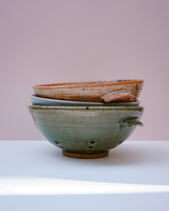 Three stacked handmade ceramic bowls with a speckled glaze, featuring a top bowl with a raw, unglazed rim, displayed on a white surface against a soft pink background.