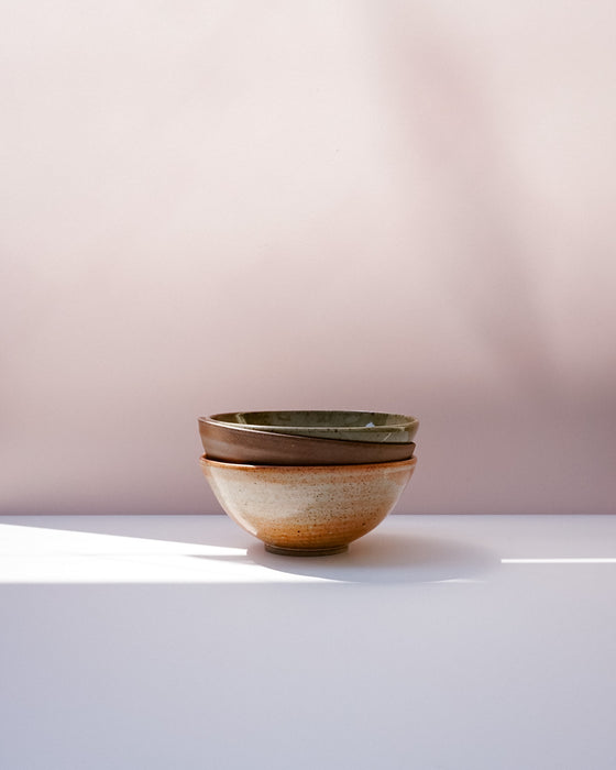Three stacked ceramic bowls with a speckled beige and brown glaze and a dark green interior, set on a white surface against a soft pink background. Morning Bowl | Scree Ceramics | Fodder