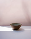 A handmade ceramic bowl with a textured brown exterior and a speckled green interior, sitting on a white surface with soft shadows against a dusty pink background. Morning Bowl | Scree Ceramics | Fodder