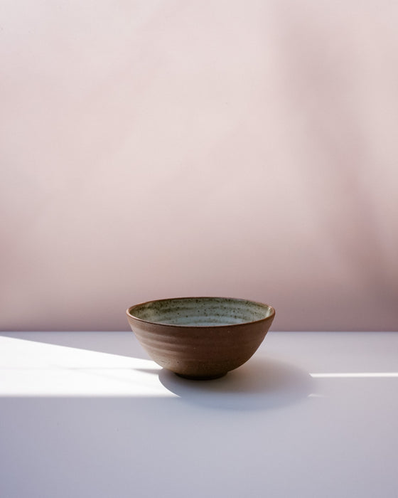 A handmade ceramic bowl with a textured brown exterior and a speckled green interior, sitting on a white surface with soft shadows against a dusty pink background. Morning Bowl | Scree Ceramics | Fodder