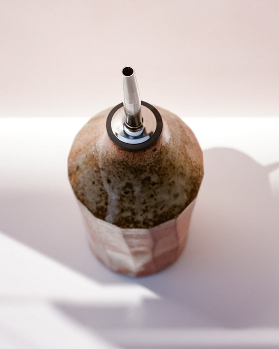 A handmade ceramic oil bottle with a rounded, textured body in earthy brown and beige glazes, featuring a metallic silver pour spout with a black rubber seal, set against a soft pink background with artistic shadows. Oil Bottle | Scree Ceramics | Fodder