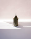 A faceted dark olive green ceramic bottle with a silver metal pour spout, standing on a white surface under dramatic diagonal sunlight. Oil Bottle | Scree Ceramics | Fodder