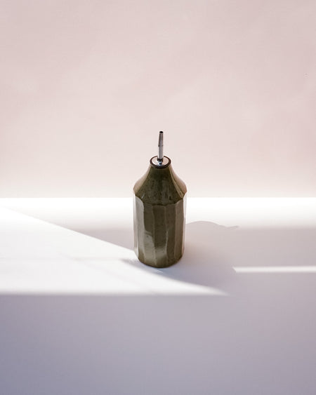 A faceted dark olive green ceramic bottle with a silver metal pour spout, standing on a white surface under dramatic diagonal sunlight. Oil Bottle | Scree Ceramics | Fodder