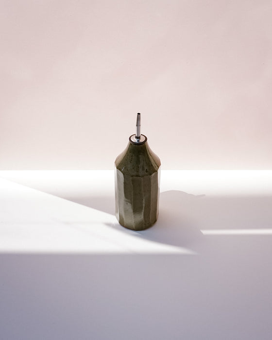 A faceted dark olive green ceramic bottle with a silver metal pour spout, standing on a white surface under dramatic diagonal sunlight. Oil Bottle | Scree Ceramics | Fodder