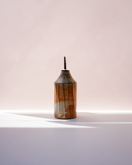 A small ceramic bottle with a conical shape, featuring a textured glaze in orange, brown, and cream, with a dark spout, set against a soft pink background with a white surface below. Oil Bottle | Scree Ceramics | Fodder