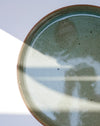 A close-up of a round ceramic plate with a speckled sage green and beige glaze, featuring a rustic terracotta rim and soft window shadow patterns across the surface | Scree Ceramics | Fodder