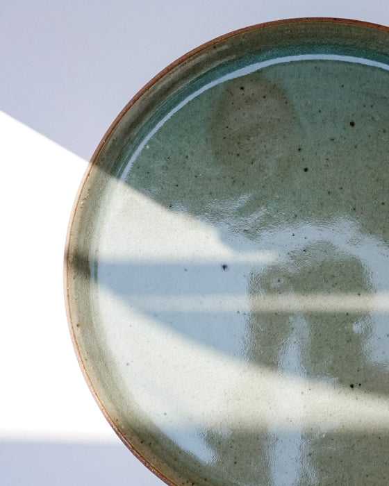 A close-up of a round ceramic plate with a speckled sage green and beige glaze, featuring a rustic terracotta rim and soft window shadow patterns across the surface | Scree Ceramics | Fodder