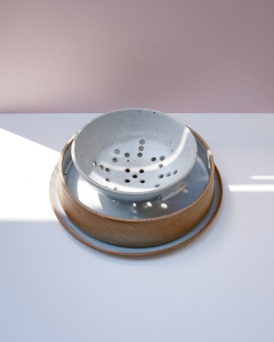 A ceramic kitchen strainer basket with a light gray speckled interior and a rustic brown glazed exterior, shown on a white surface with a soft pink background | Scree Ceramics | Fodder