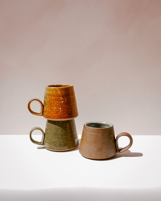 Three handmade ceramic mugs with organic shapes and handles, stacked and standing on a white surface against a warm beige background. Tall Loop Mug | Scree Ceramics | Fodder