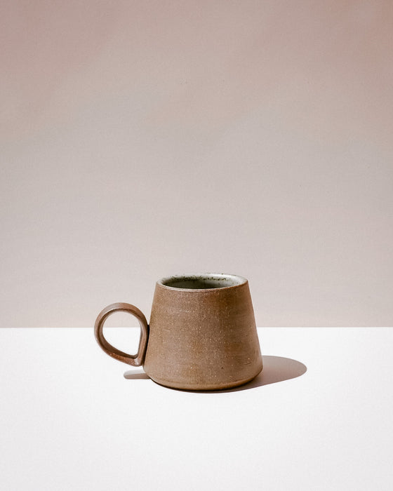 A small handmade ceramic mug with a tapered shape and a loop handle, featuring a textured earthy brown exterior and a dark green glazed interior, set against a minimalist beige and white background. Tall Loop Mug | Scree Ceramics | Fodder