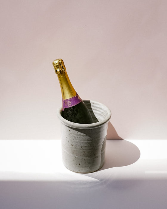 A sparkling wine bottle with a gold foil top and purple neck label, resting in a speckled gray ceramic bucket on a white surface against a soft pink background. Scree Ceramics | Fodder