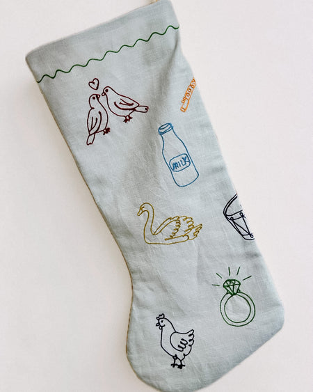 A light gray fabric Christmas stocking with embroidered holiday-themed illustrations including birds, a milk bottle, a swan, a glass, a chicken, and a ring, shown on a plain white background.12 Days of Christmas embroidered linen Christmas stocking by See & Quin - carol motifs - Fodder