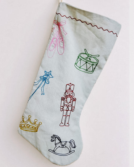 A light blue fabric Christmas stocking decorated with colorful embroidered toy-themed motifs including a nutcracker, drum, ballet shoes, wand, crown, and rocking horse, shown against a plain off-white background. Nutcracker embroidered linen Christmas stocking by See & Quin - ballet motifs - Fodder