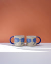 Two ceramic mugs with blue floral designs on a white surface and orange background. Sophie Alda | Handmade Ceramics | Shop Fodder