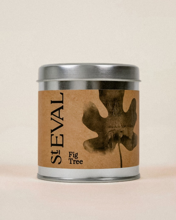 A cylindrical metal tin candle with a silver lid and base, featuring a brown kraft paper label with a dark brown fig tree leaf graphic and the text 'St EVAL Fig Tree'.