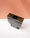 A modern sculptural metal container or candle holder with a rectangular top, a central square opening, and two side slots, shown on a white surface against a peach-colored background. Studio ERS | Soup Vase | Fodder