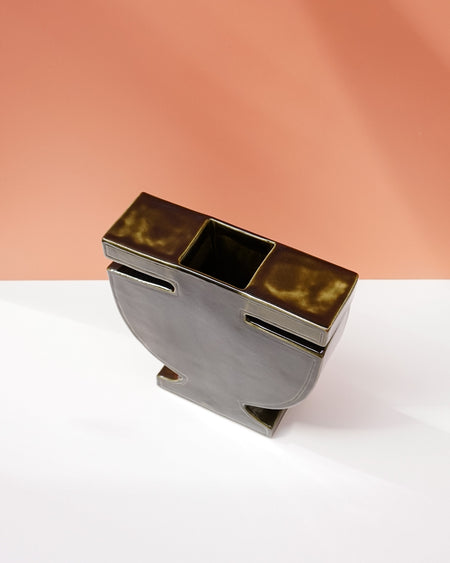 A modern sculptural metal container or candle holder with a rectangular top, a central square opening, and two side slots, shown on a white surface against a peach-colored background. Studio ERS | Soup Vase | Fodder