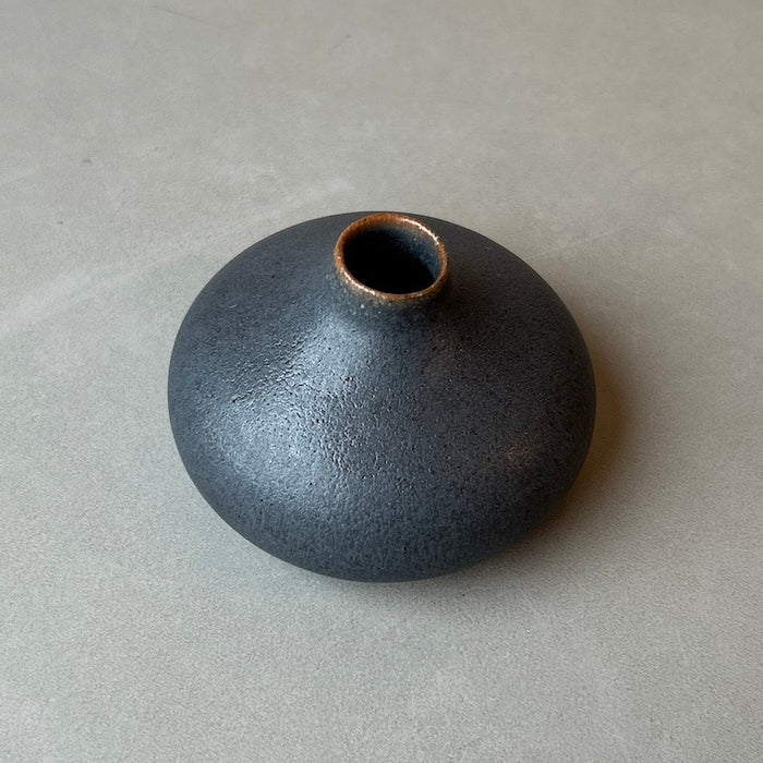 a dark charcoal coloured bud vase on a light grey table. 